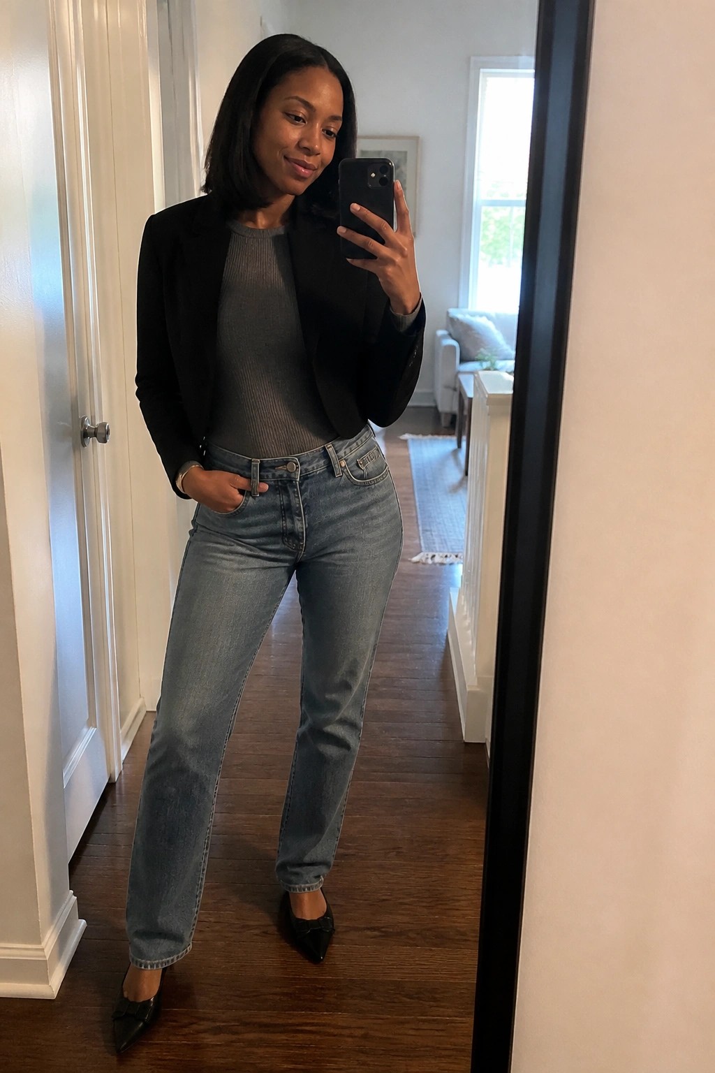 Woman in black blazer and jeans taking mirror selfie with smartphone in hallway