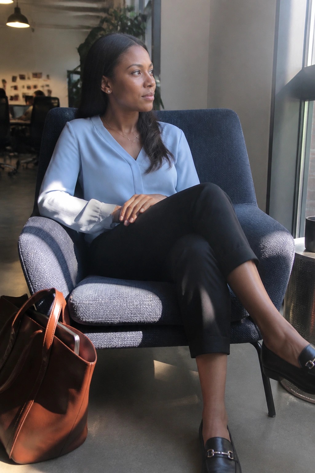 Woman in light blue blouse and black pants seated in blue armchair beside brown bag.