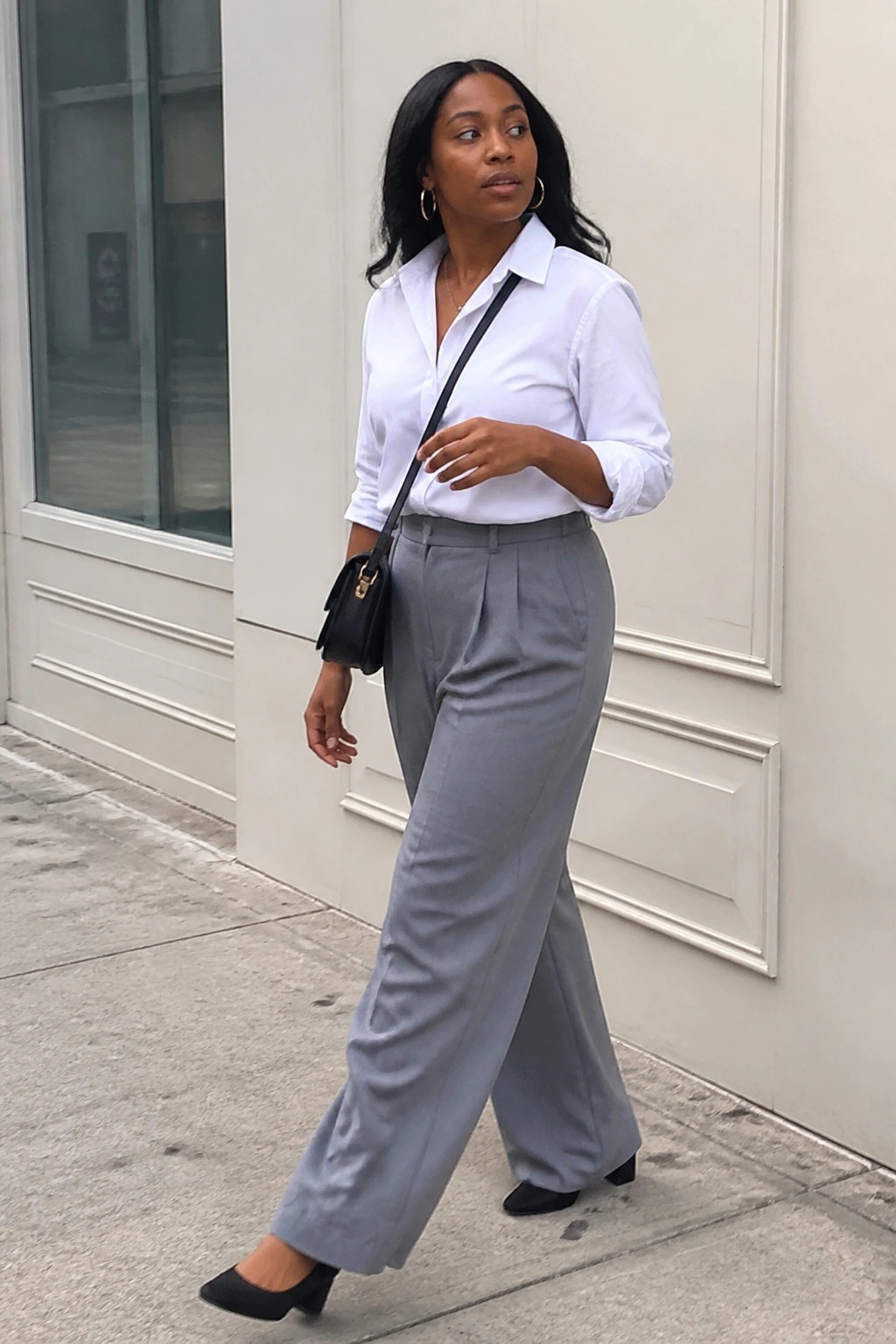 Woman in white shirt and gray wide-leg pants walking on sidewalk with black bag