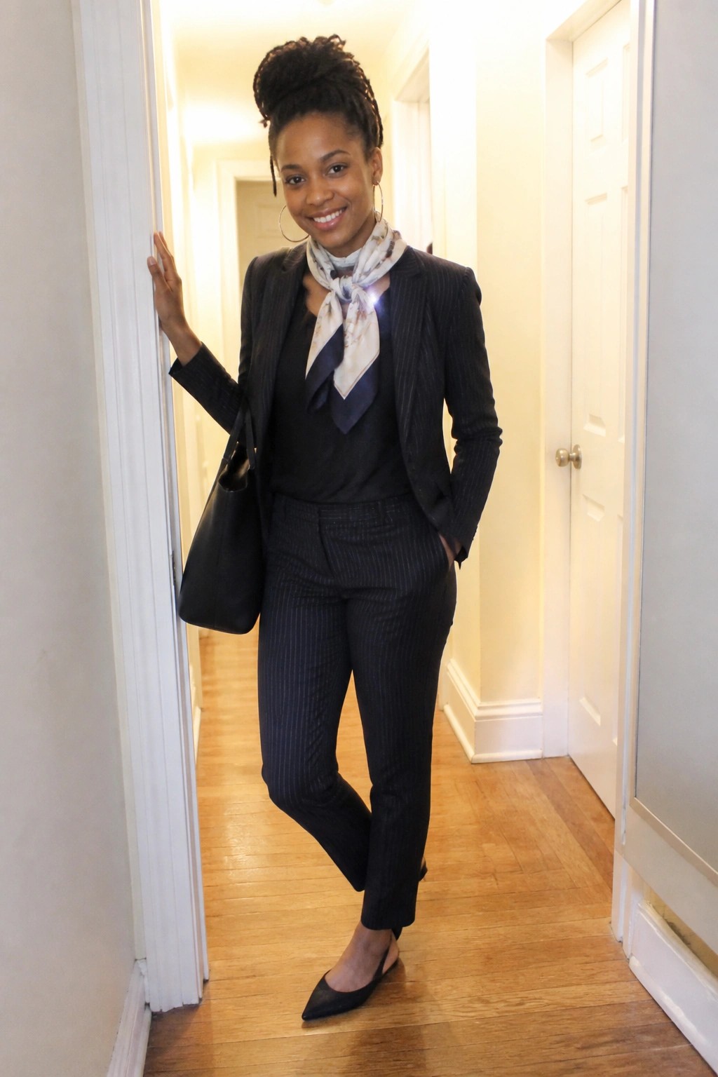 Smiling woman in pinstripe suit and scarf posing in hallway with handbag.