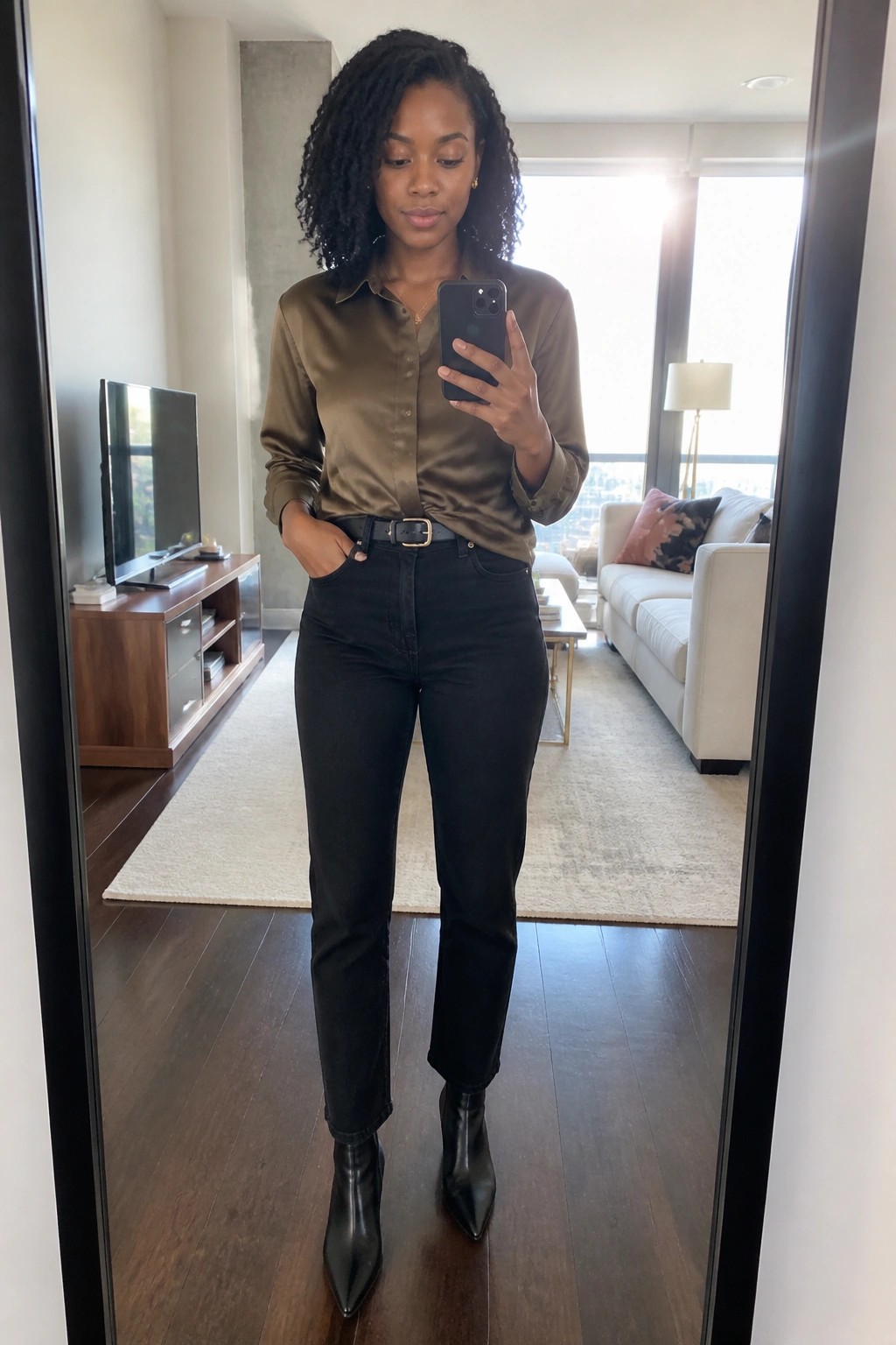 Black woman wearing an olive satin button-up shirt tucked into black jeans with a black belt and pointed black ankle boots while taking a mirror selfie.