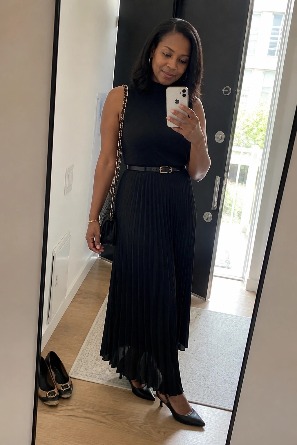 Woman wearing a black sleeveless mock neck top tucked into a black pleated midi skirt with a thin black belt, black pointed toe heels, and a black quilted handbag with gold chain strap.