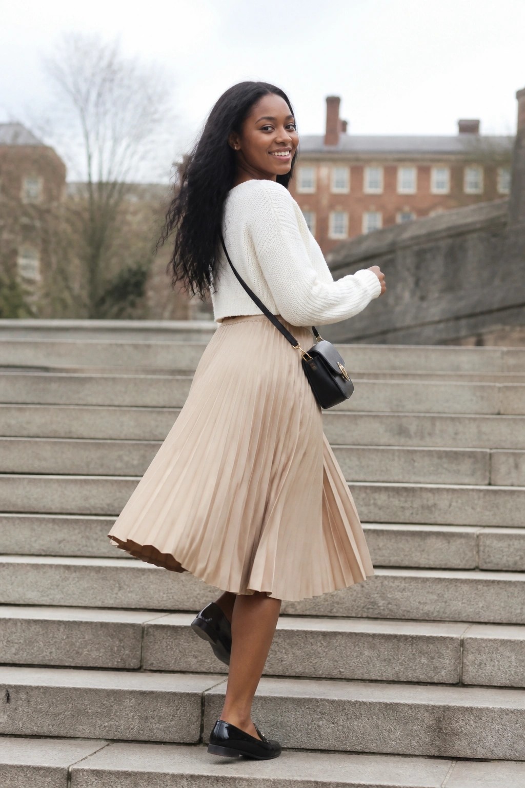 Woman in white sweater and beige pleated skirt on stone steps with black bag