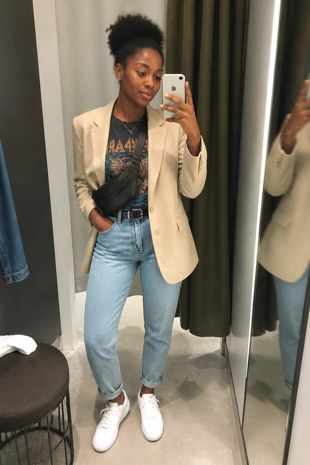 Woman in beige blazer and blue jeans taking iPhone mirror selfie in fitting room