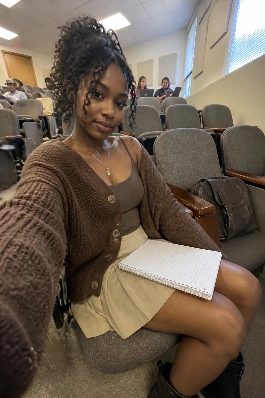 Curly-haired woman in brown cardigan takes selfie in lecture hall with notebook.