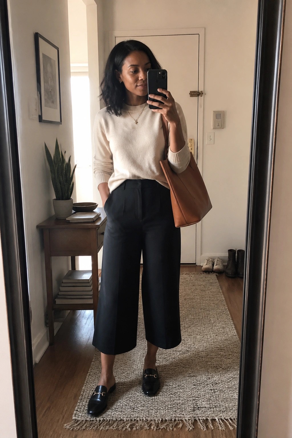 Woman in cream knit sweater, black wide cropped pants, black loafers, and brown leather tote taking a mirror selfie in a hallway.