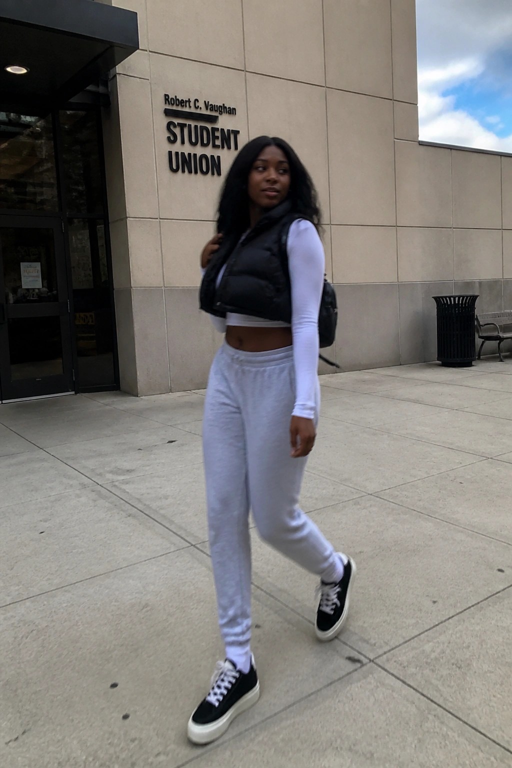 Black woman wearing a white long sleeve crop top under a black puffer vest, light gray sweatpants, and black platform sneakers while carrying a backpack outside a student union building.