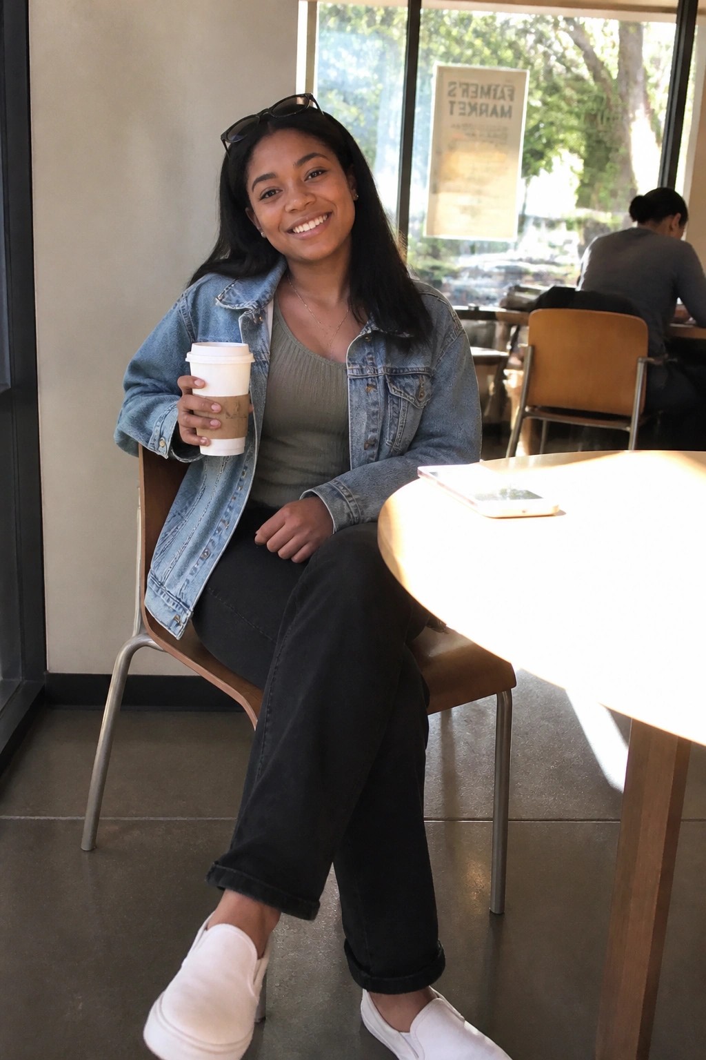 Smiling young woman in denim jacket holds coffee cup in sunny cafe.