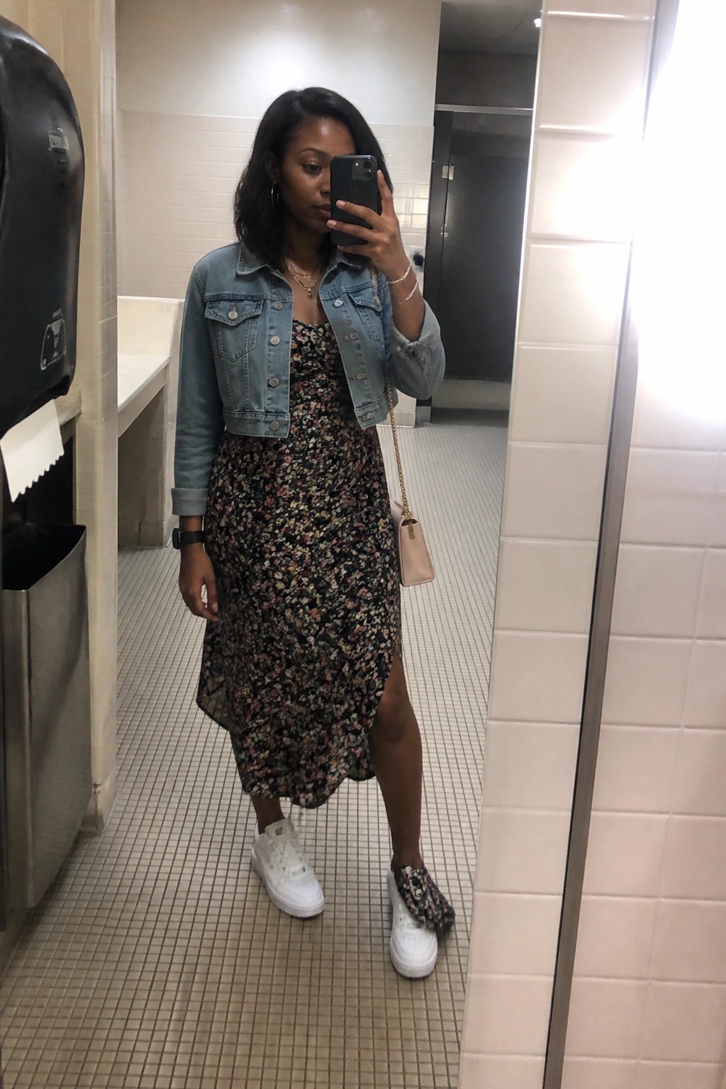Woman in floral midi dress and denim jacket taking mirror selfie in restroom