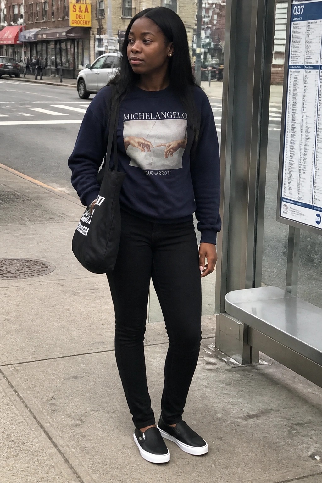 Woman in navy Michelangelo sweatshirt and black jeans at bus stop