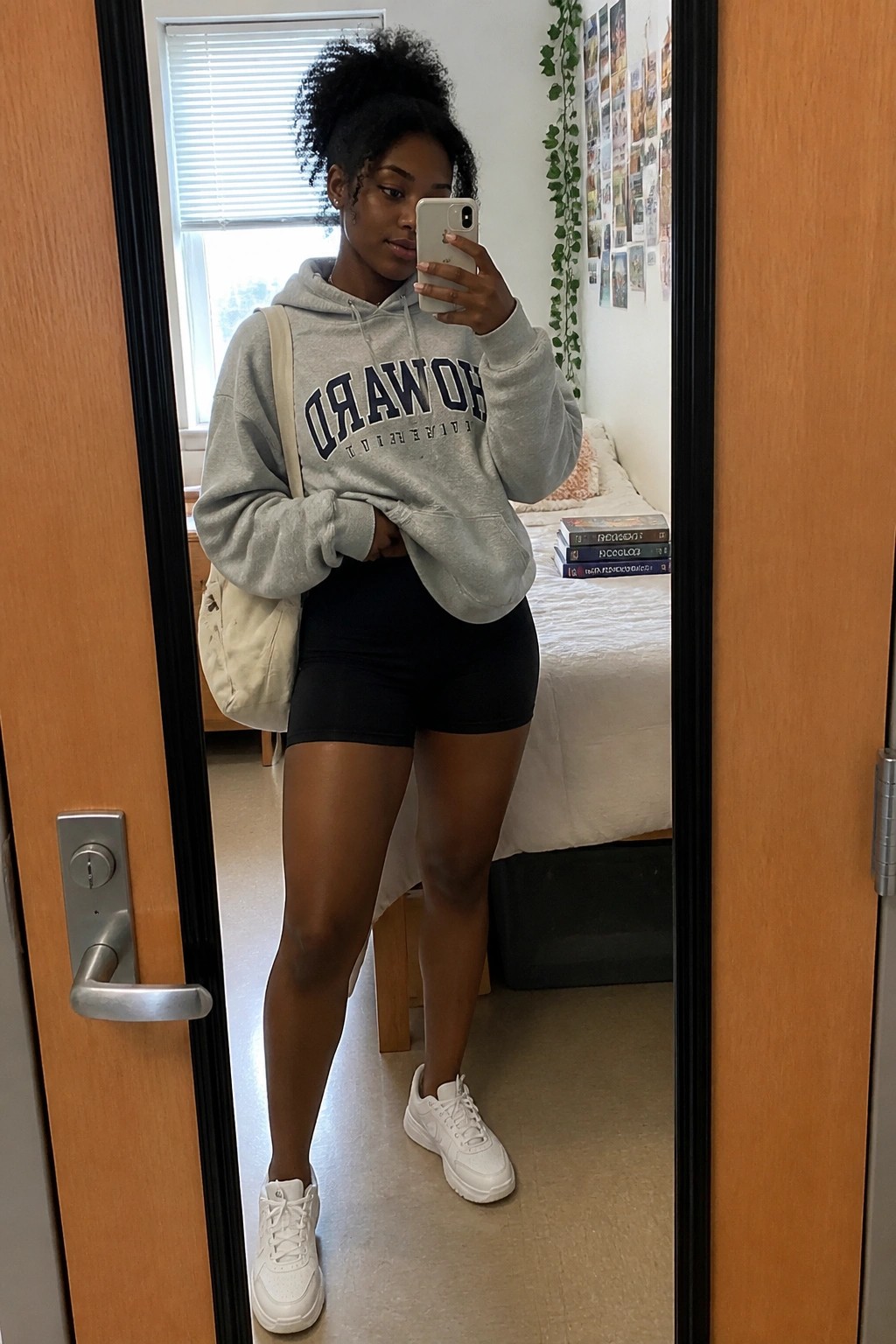 A young Black woman taking a mirror selfie in an oversized grey Howard University hoodie, black biker shorts, white sneakers, and a white tote bag slung over one shoulder.