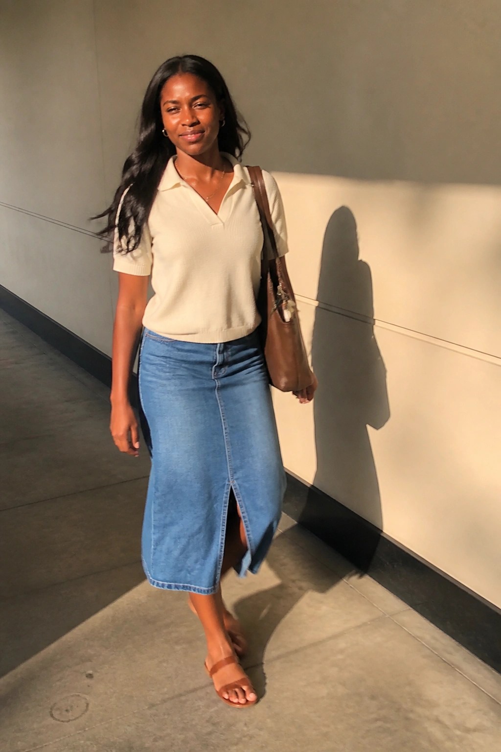 Woman in cream polo sweater and blue denim skirt with brown tote bag in hallway