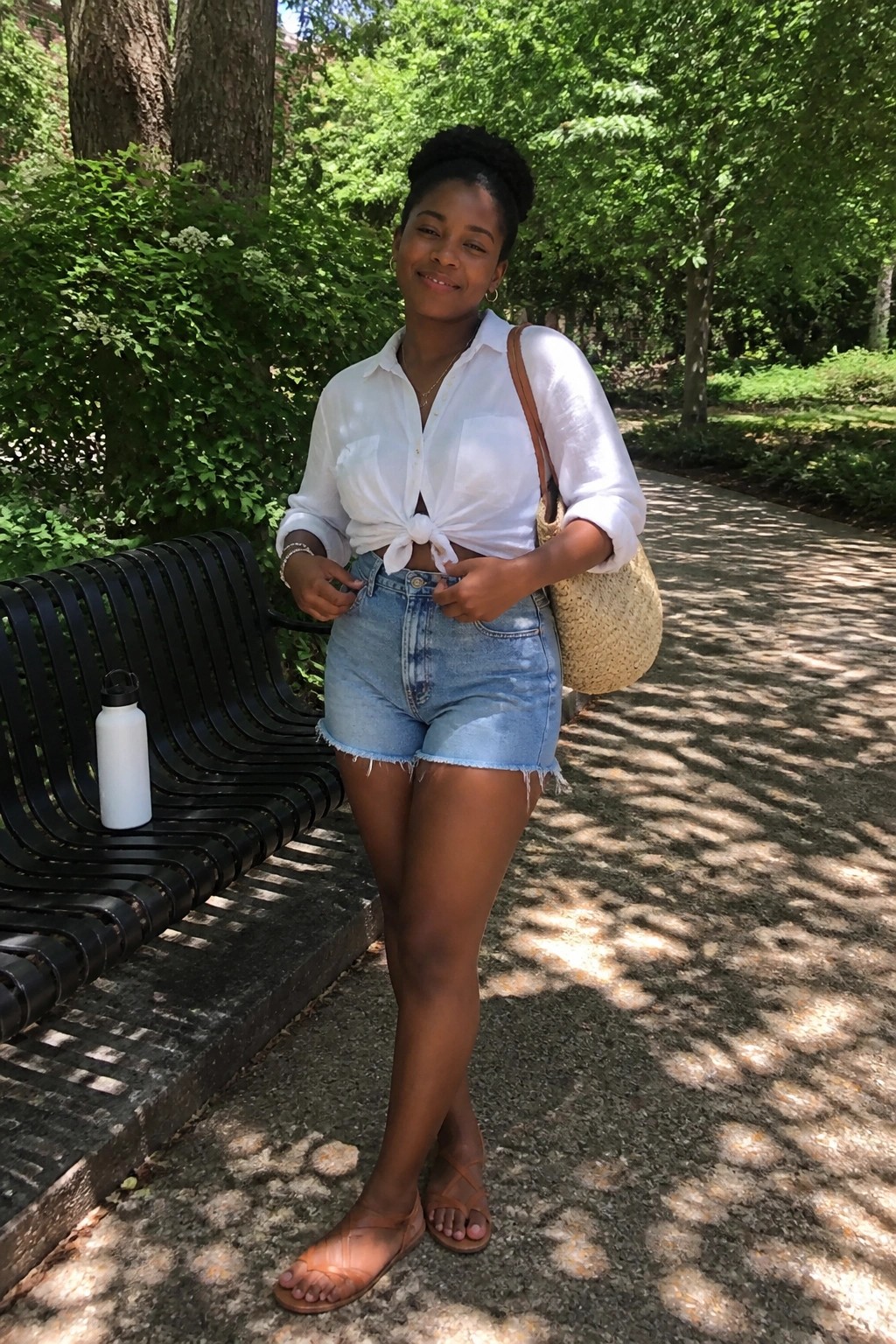 Smiling woman in tied white shirt and denim shorts near black bench in park.