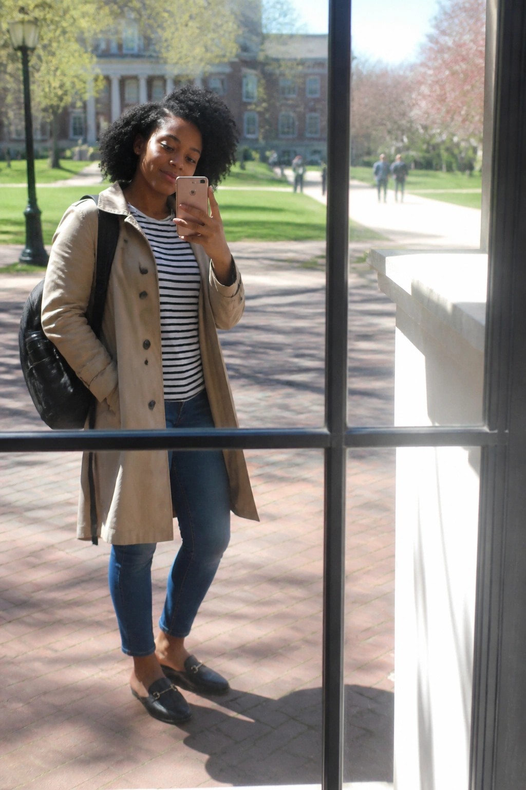 Woman in beige trench coat taking selfie on sunny brick campus path