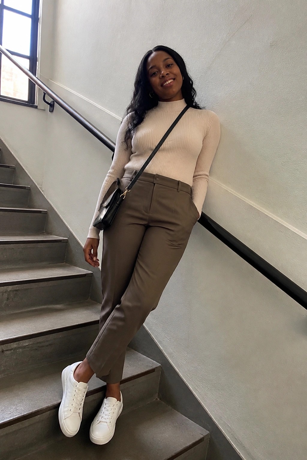 Woman wearing a cream ribbed turtleneck sweater tucked into taupe straight-leg trousers, white sneakers, and a small black crossbody bag while standing on indoor stairs.