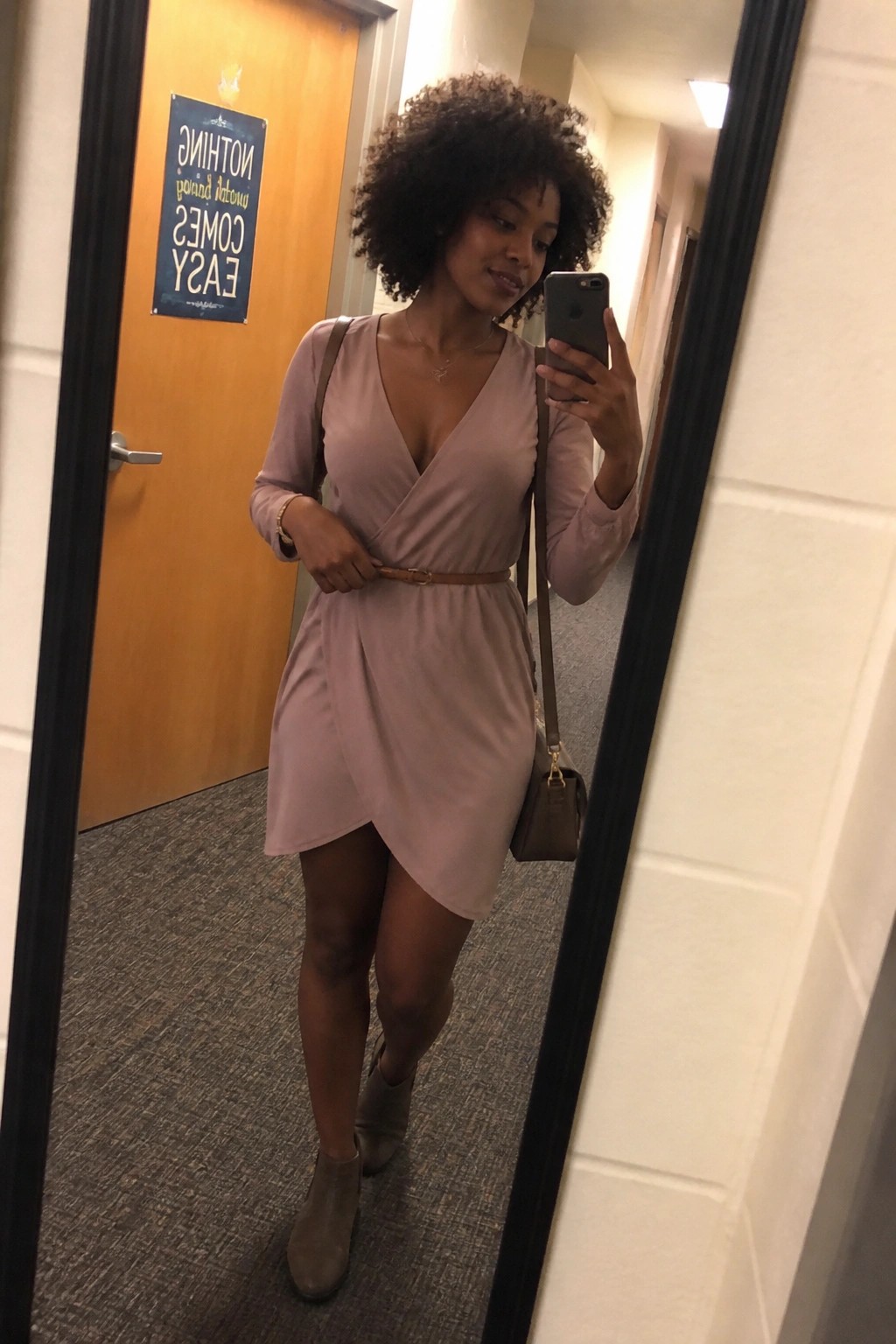 Woman with curly hair in pink wrap dress taking mirror selfie in hallway with boots