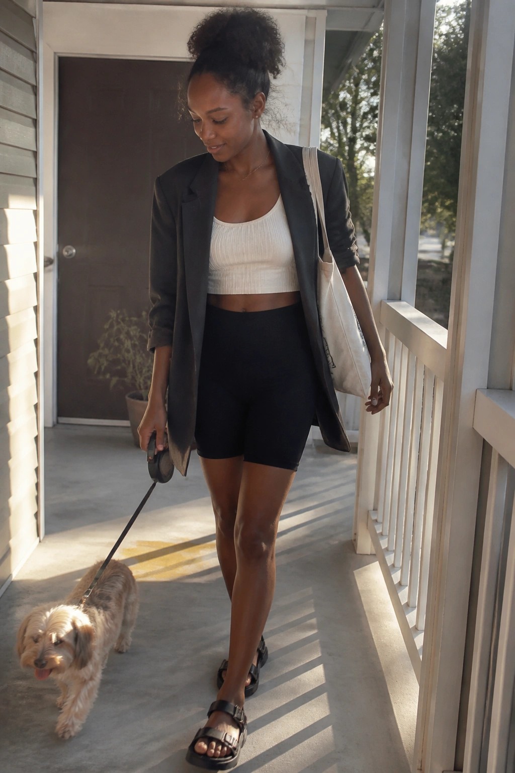 Woman in black blazer and shorts walks small dog on sunny porch with tote bag.