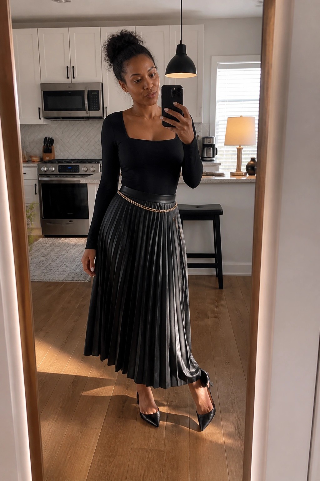 Woman wearing a black square neck bodysuit tucked into a pleated black midi skirt with gold chain belt and black pointed toe heels while taking a mirror selfie.