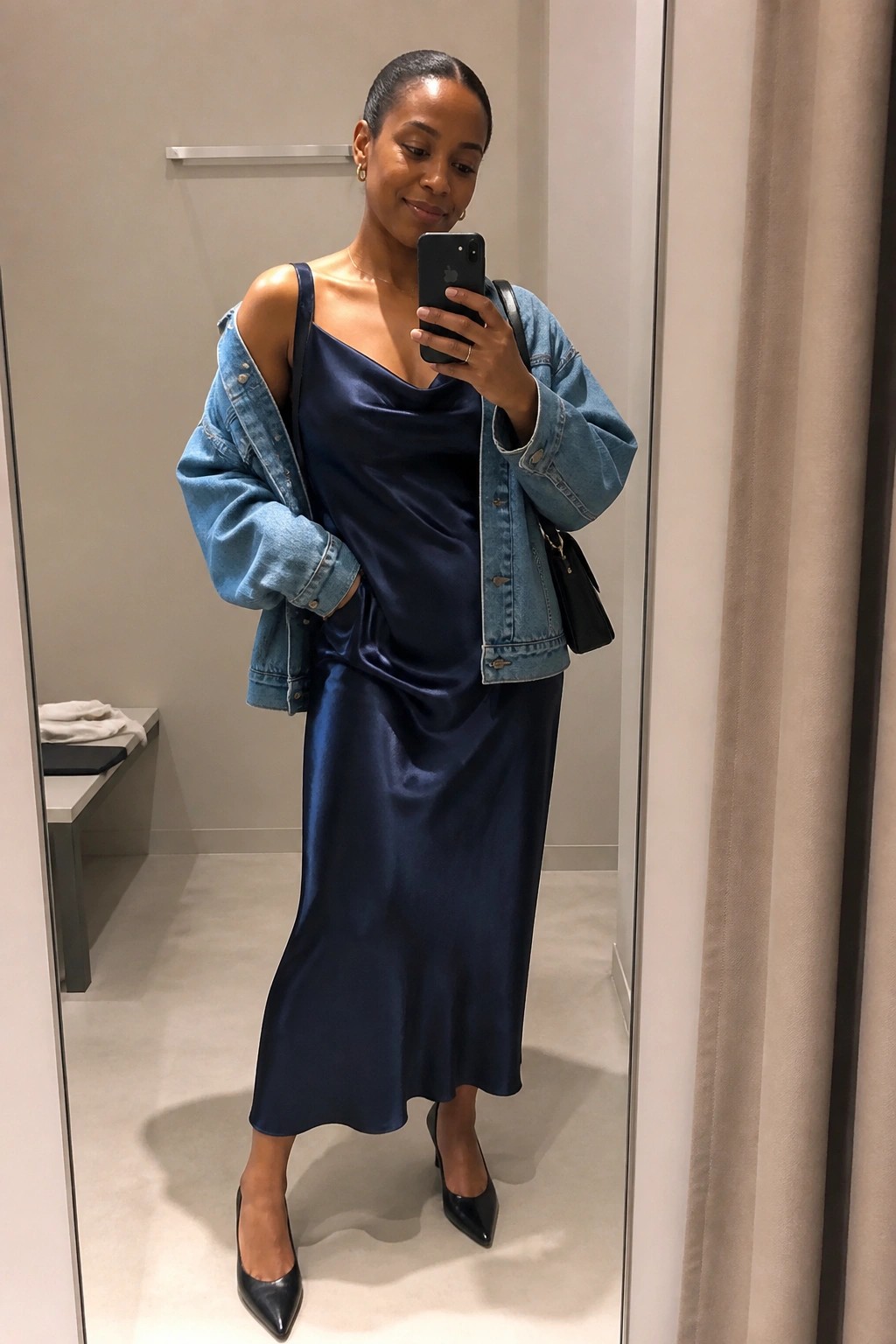 Woman wearing a navy satin midi slip dress layered with an oversized blue denim jacket, black pointed toe heels, and a small black handbag while taking a mirror selfie.