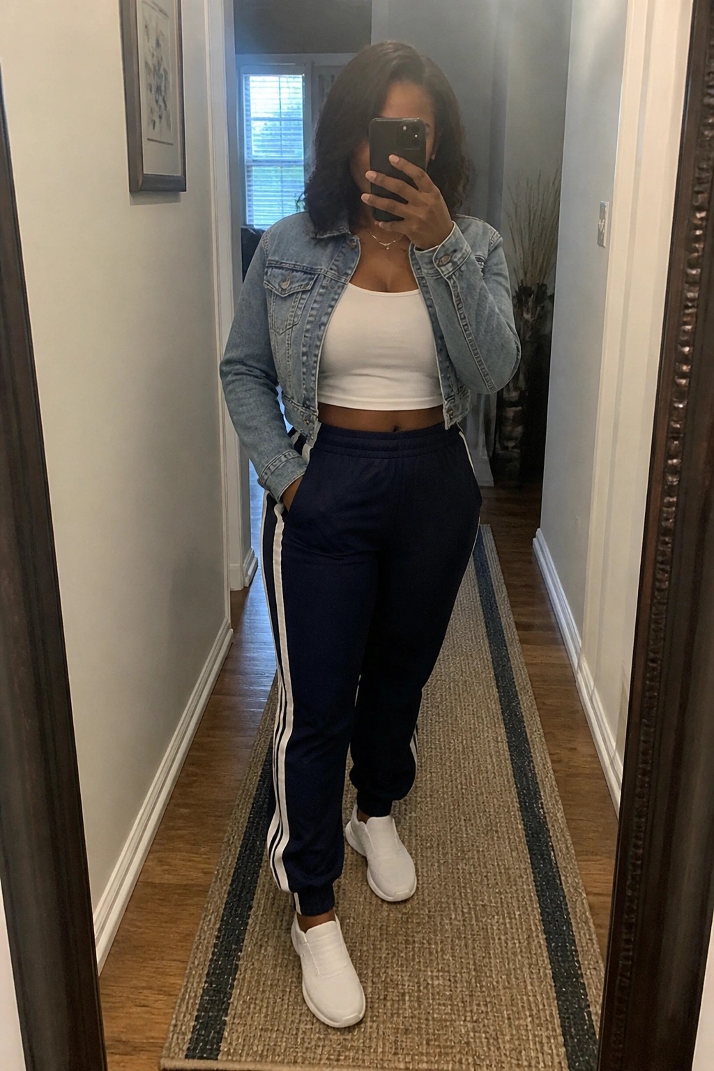 Woman in denim jacket, white crop top, navy track pants taking mirror selfie