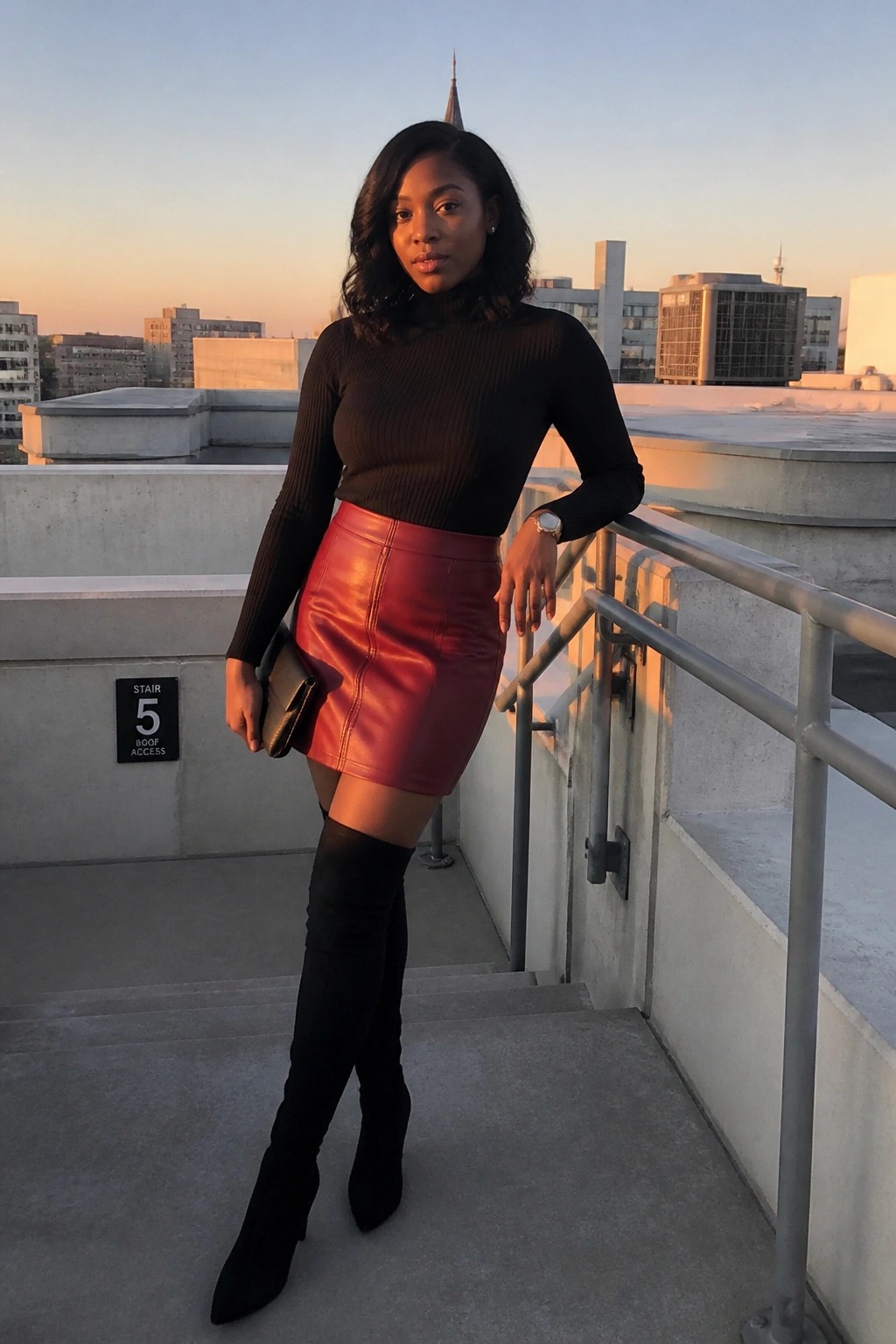 Woman in black ribbed turtleneck tucked into red leather mini skirt with black over the knee boots holding a small black clutch on stairs.