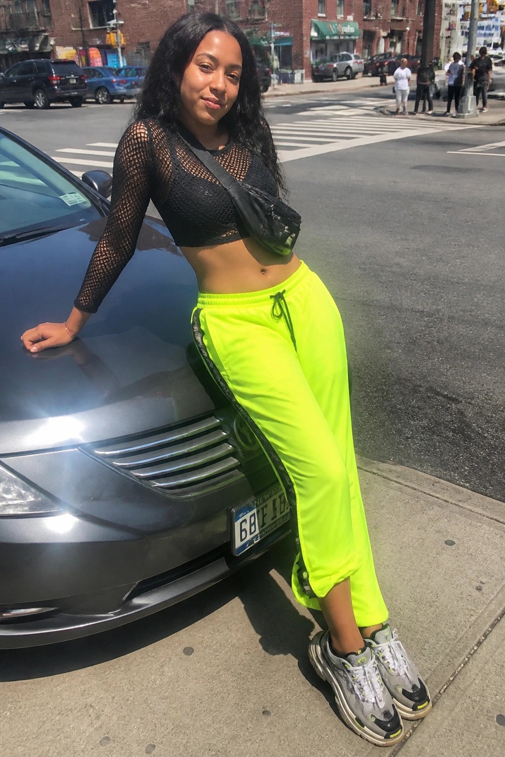 Woman in black mesh crop top, neon yellow track pants with side stripes, black crossbody bag, and gray sneakers leaning against a dark car on a city street.