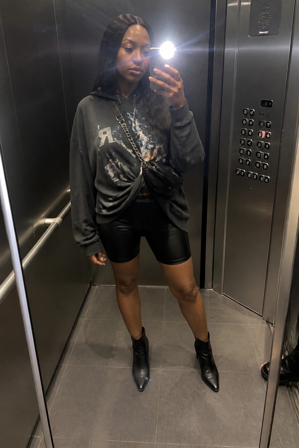 Woman taking mirror selfie in elevator wearing gray hoodie and black shorts.