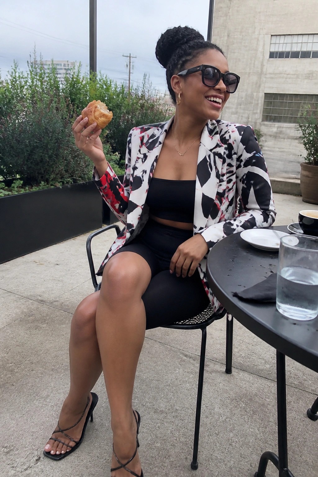 Woman seated outdoors wearing a black white and red patterned blazer over a black crop top and black shorts with strappy black heels and gold jewelry while holding a pastry.
