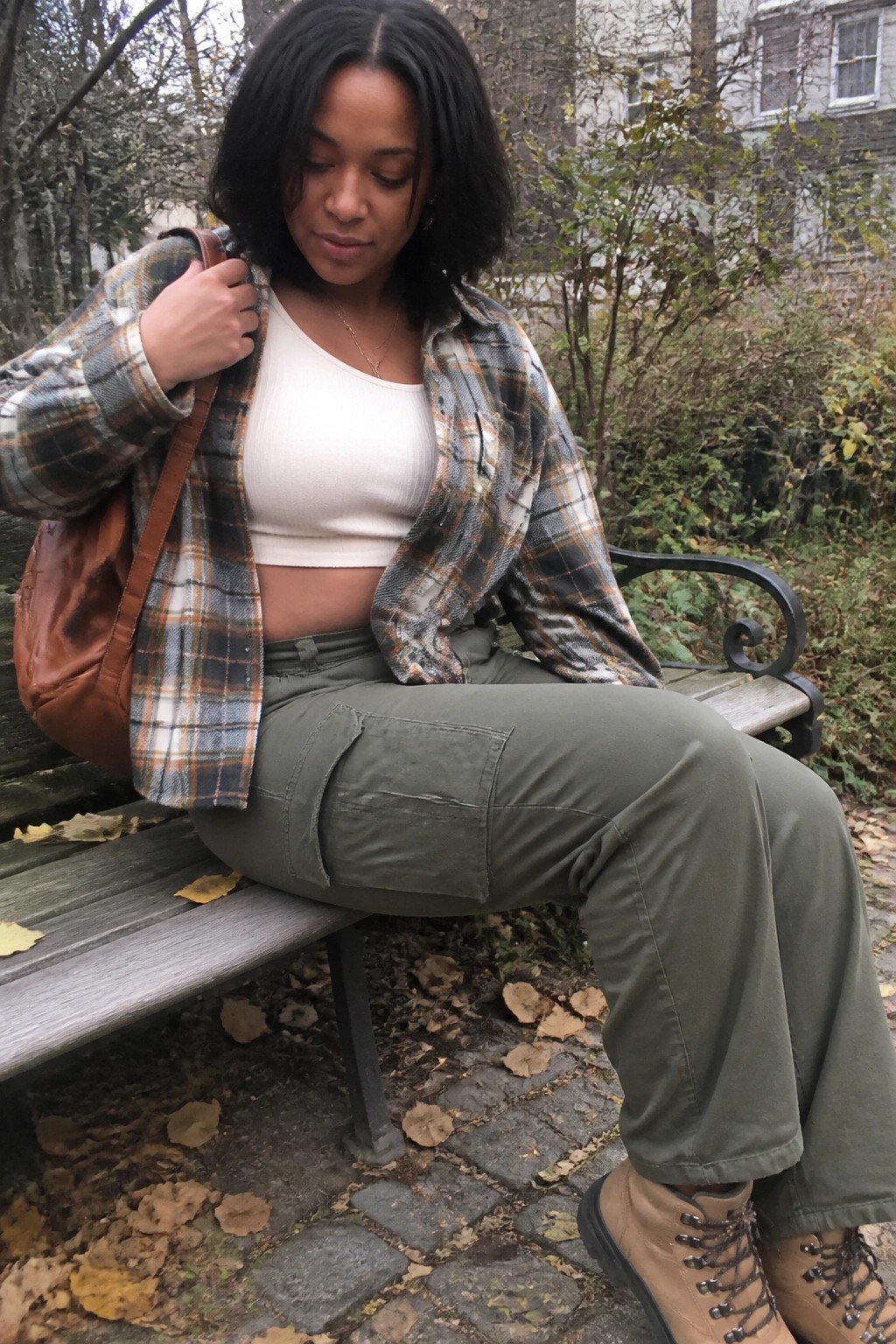 Woman in open plaid shirt and green cargo pants sits on autumn park bench with brown bag.