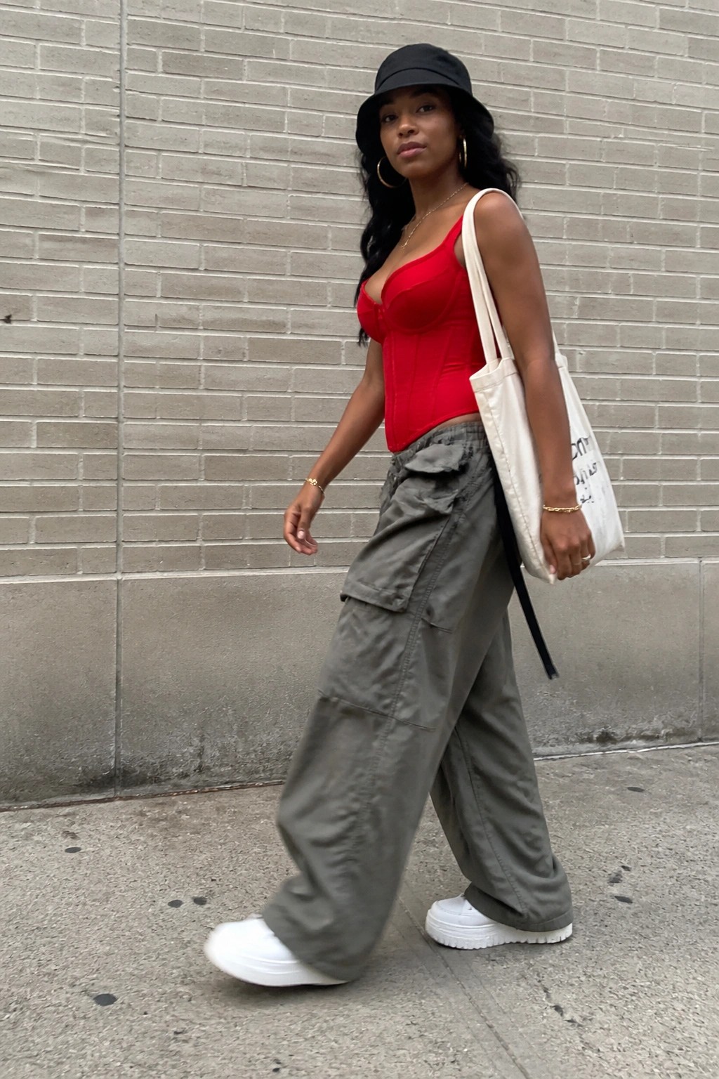 Woman wearing red corset top grey cargo pants white platform sneakers black bucket hat and gold jewelry while carrying white tote bag on city sidewalk
