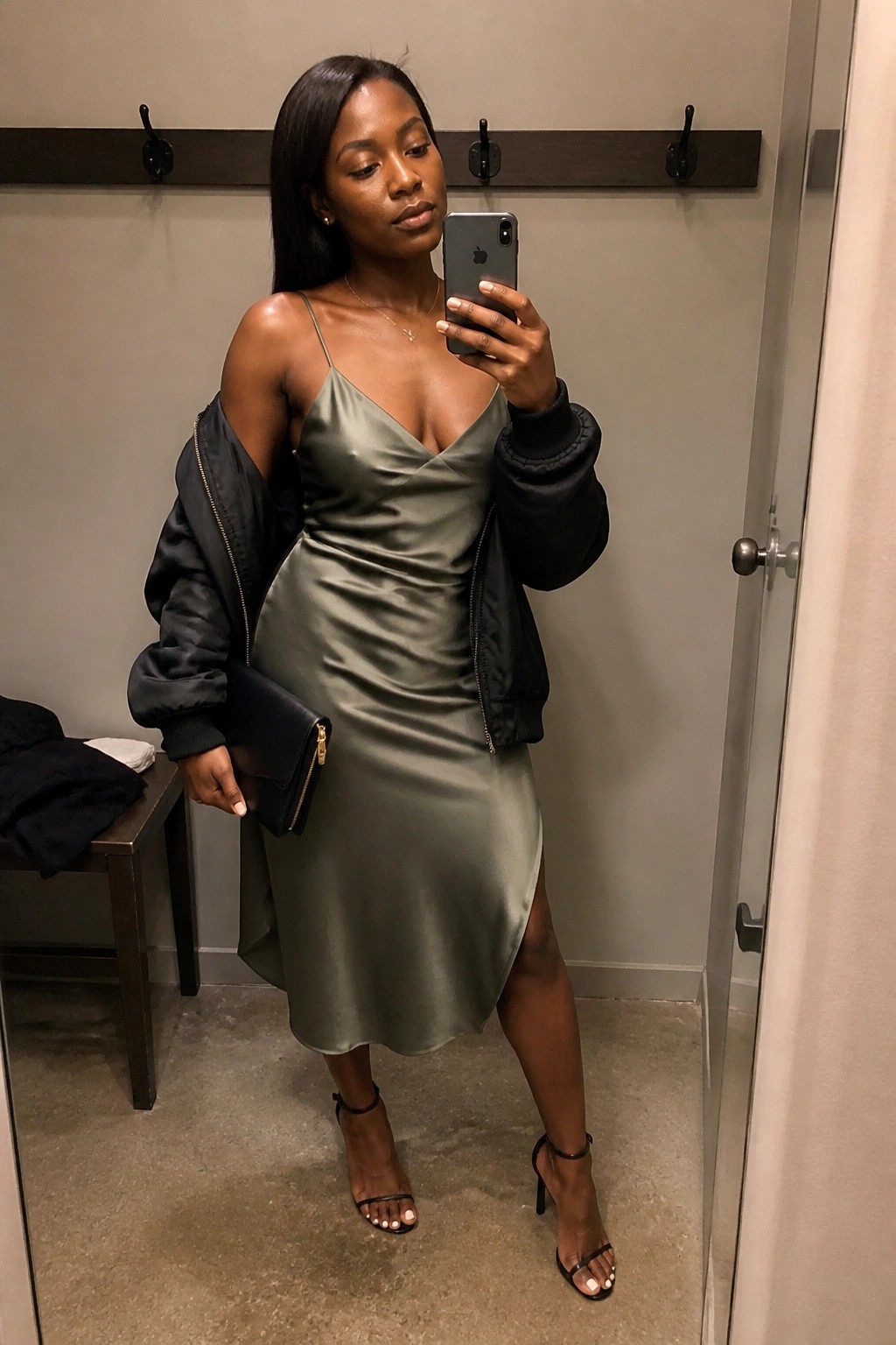 Woman in sage green satin dress with black bomber jacket taking mirror selfie.