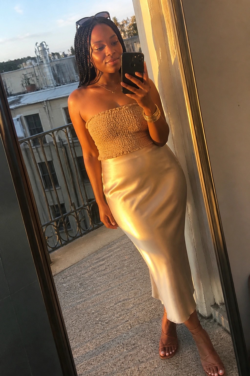 Woman wearing beige textured strapless top with beige satin midi skirt, brown strappy heels, and gold jewelry while holding a phone for a mirror selfie.