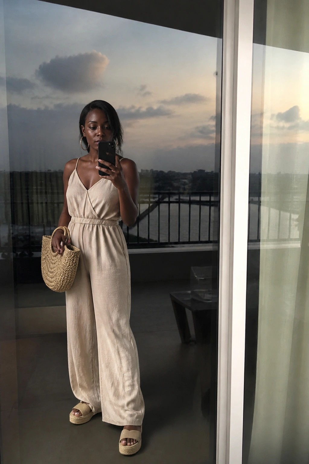 Woman wearing a beige wide leg jumpsuit with spaghetti straps and a woven straw bag while holding a phone in a mirror selfie.