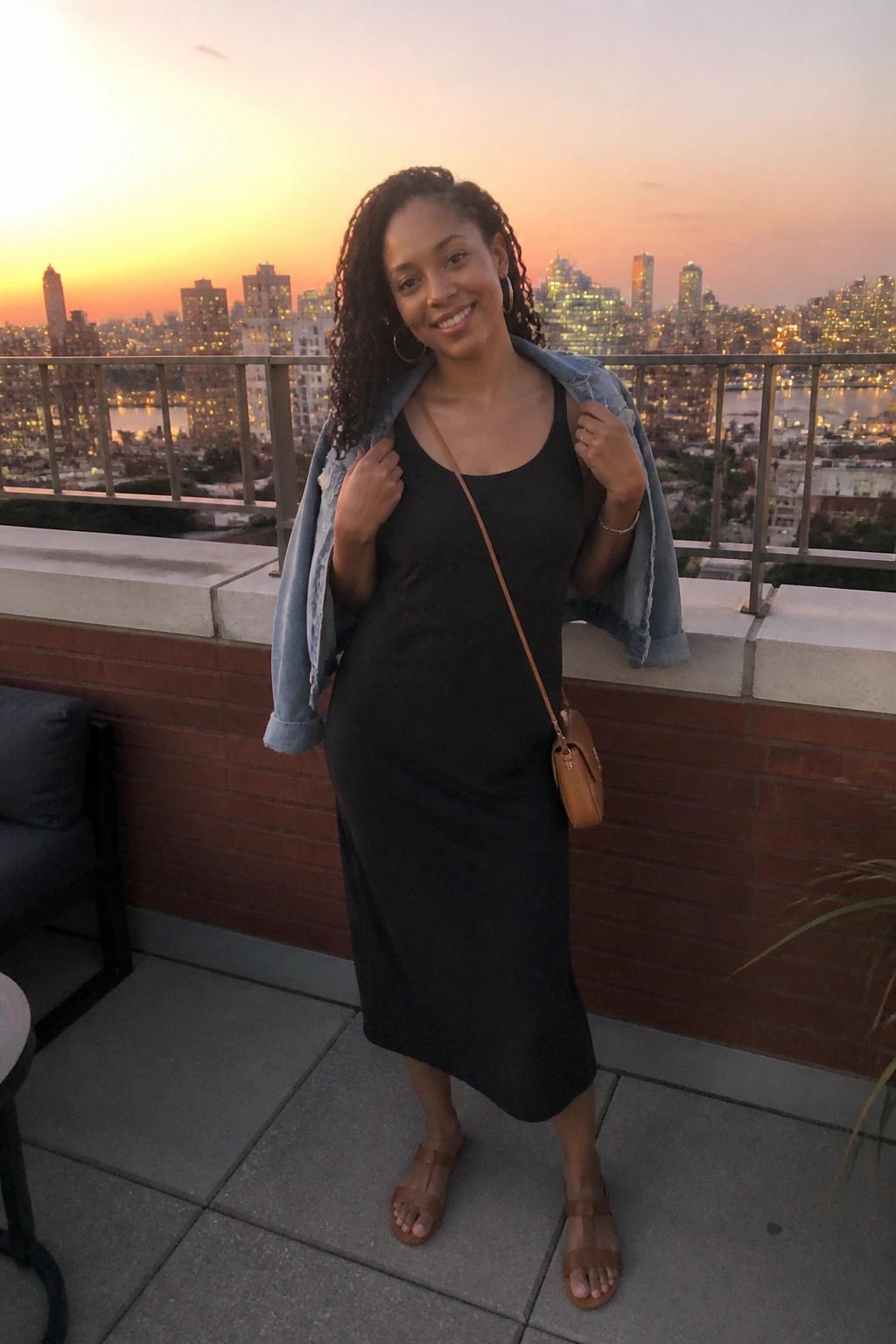 Smiling woman in black dress with denim jacket on rooftop overlooking city at sunset