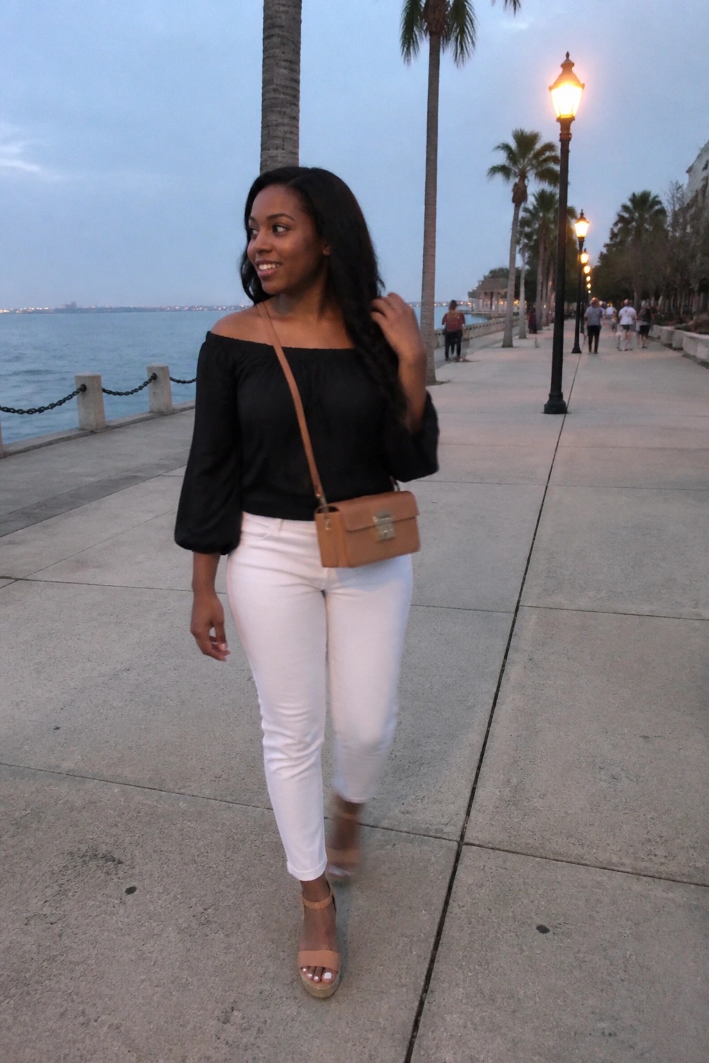 Woman in black off-shoulder top and white pants on palm-lined waterfront promenade