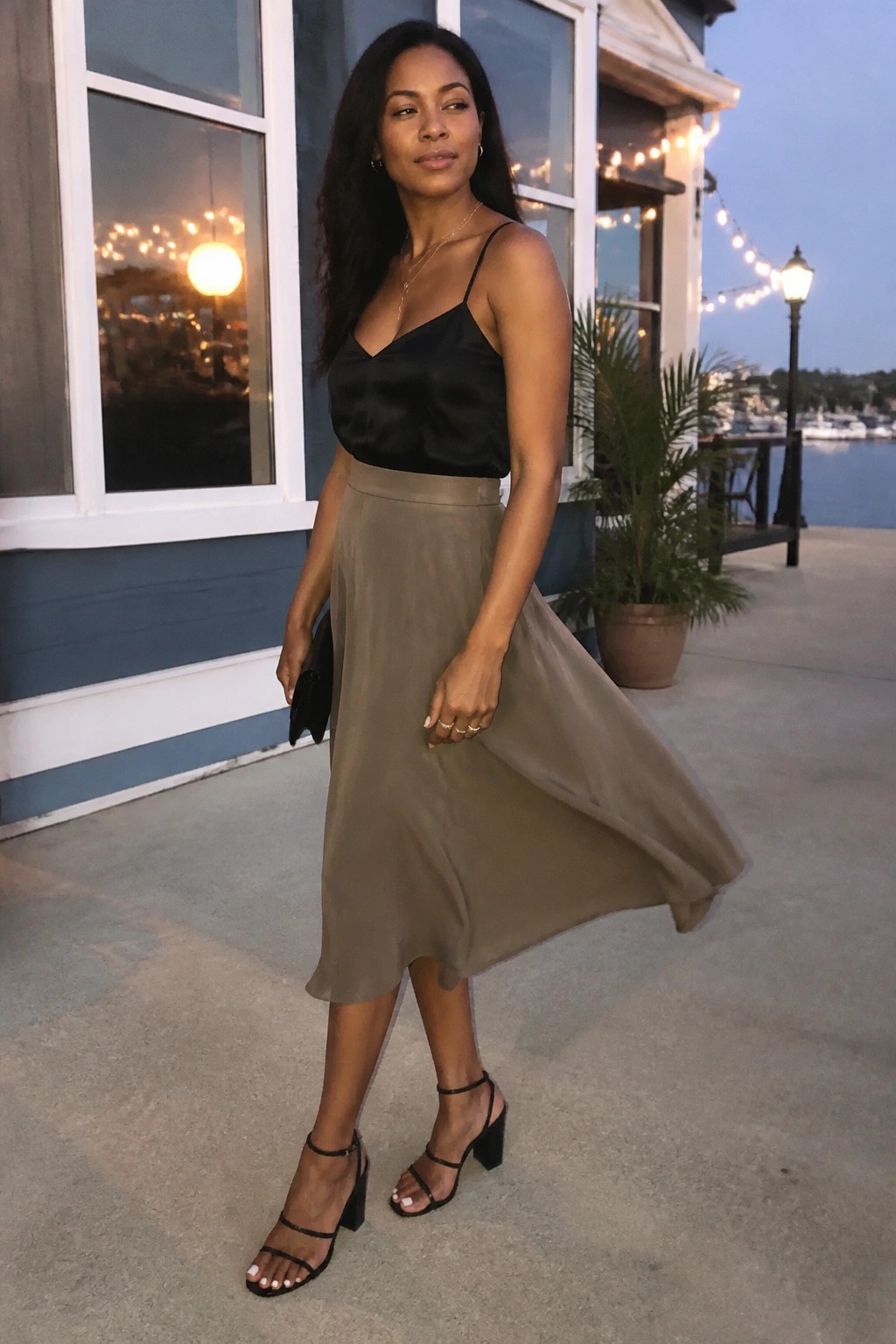 Woman wearing black satin camisole top, flowing taupe midi skirt, black strappy block heel sandals, and gold necklaces while holding a small black clutch.