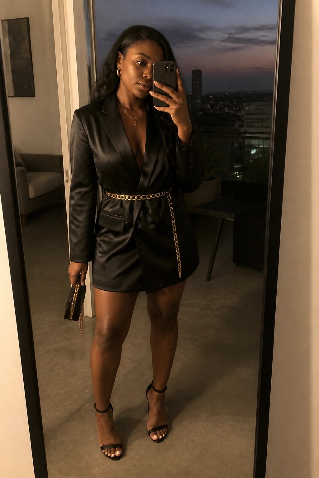 Woman in black satin blazer dress with gold chain belt taking mirror selfie at dusk.