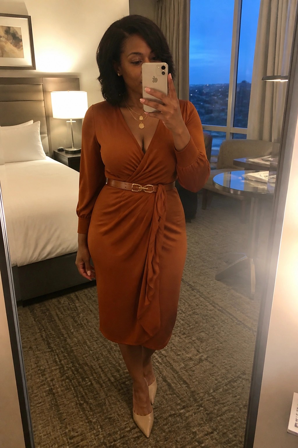Woman in a burnt orange satin wrap dress with long sleeves, brown belt with gold buckle, layered gold necklaces, and beige pointed toe heels taking a mirror selfie in a hotel room.