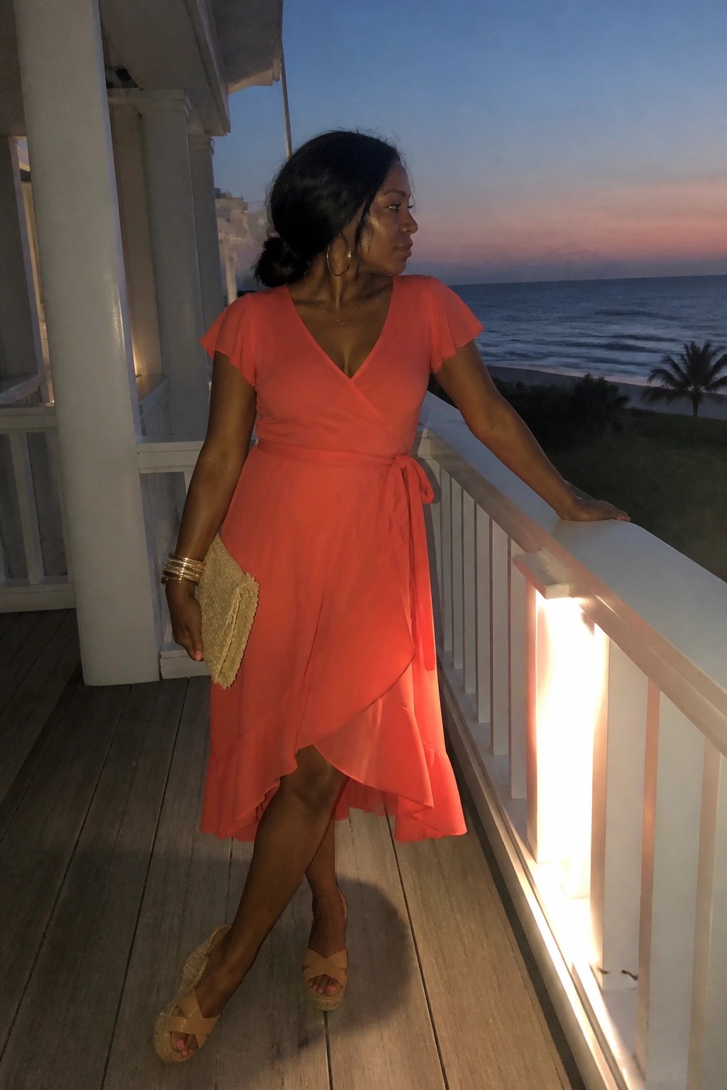 Woman in coral wrap dress on oceanfront deck at dusk holding clutch