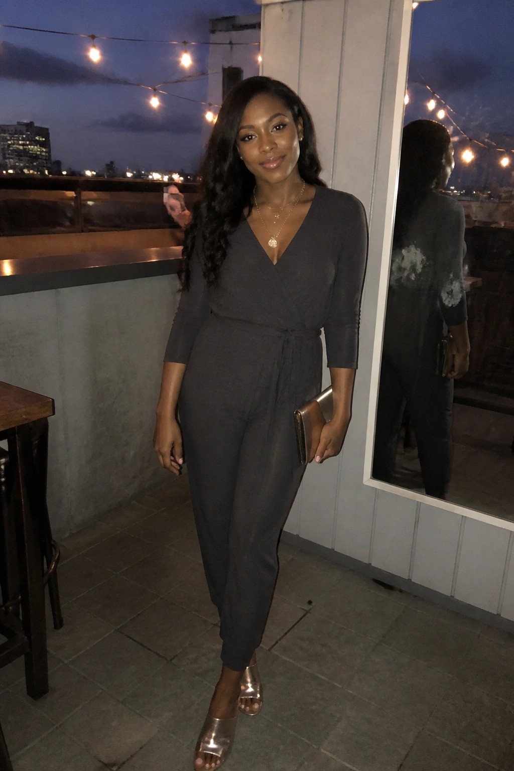 Woman in dark gray jumpsuit posing on rooftop at night with string lights
