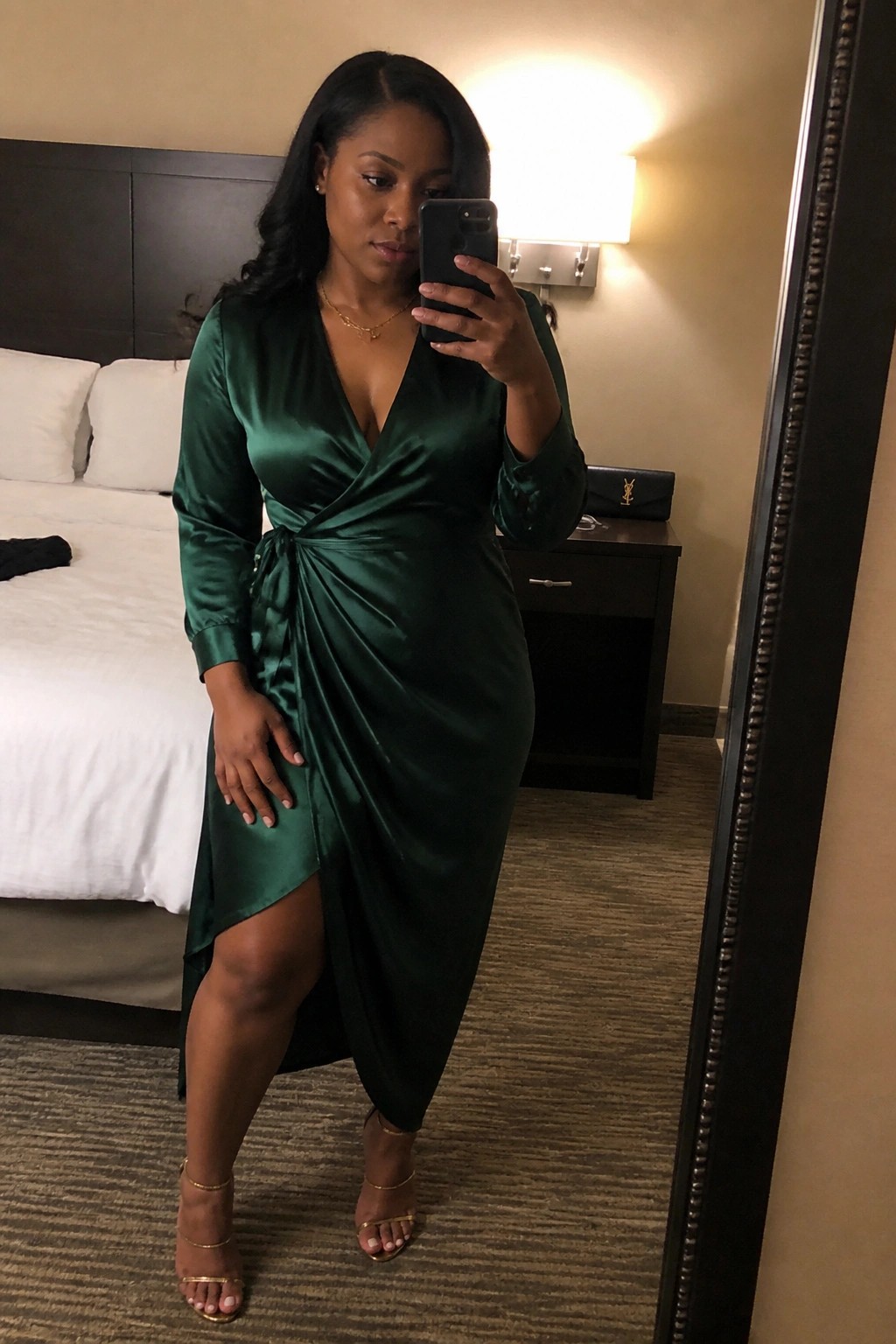 Plus size woman wearing a deep green satin wrap midi dress with long sleeves, ruched waist tie, and side slit, paired with gold strappy heels and delicate gold jewelry while holding a phone for a mirror selfie.