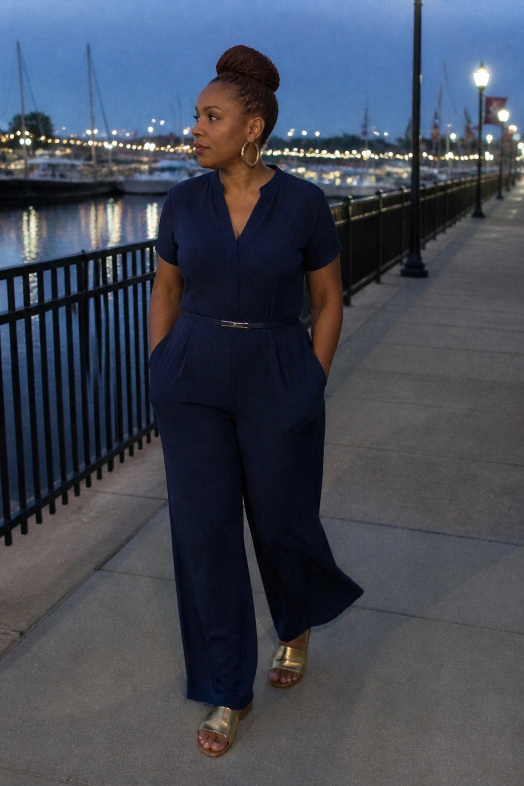 Woman wearing a navy short sleeve jumpsuit with a thin metallic belt, wide legs, and gold slide sandals paired with large gold hoop earrings.