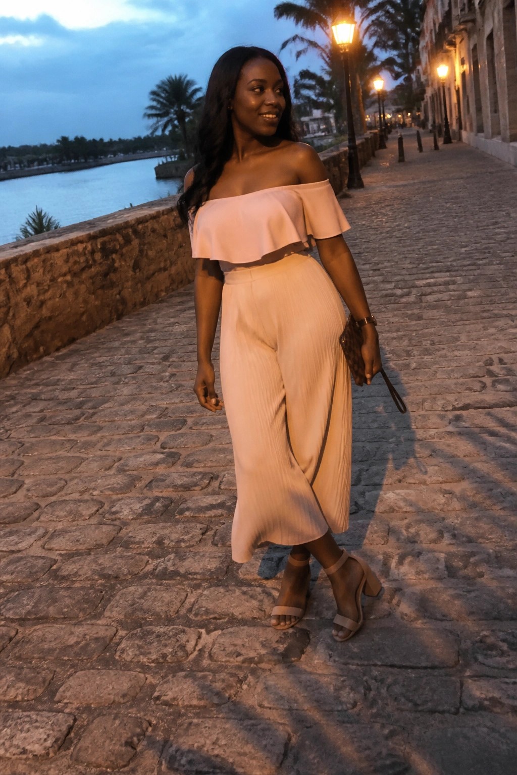 Woman in light pink off-shoulder top and pleated pants on cobblestone waterfront at dusk.