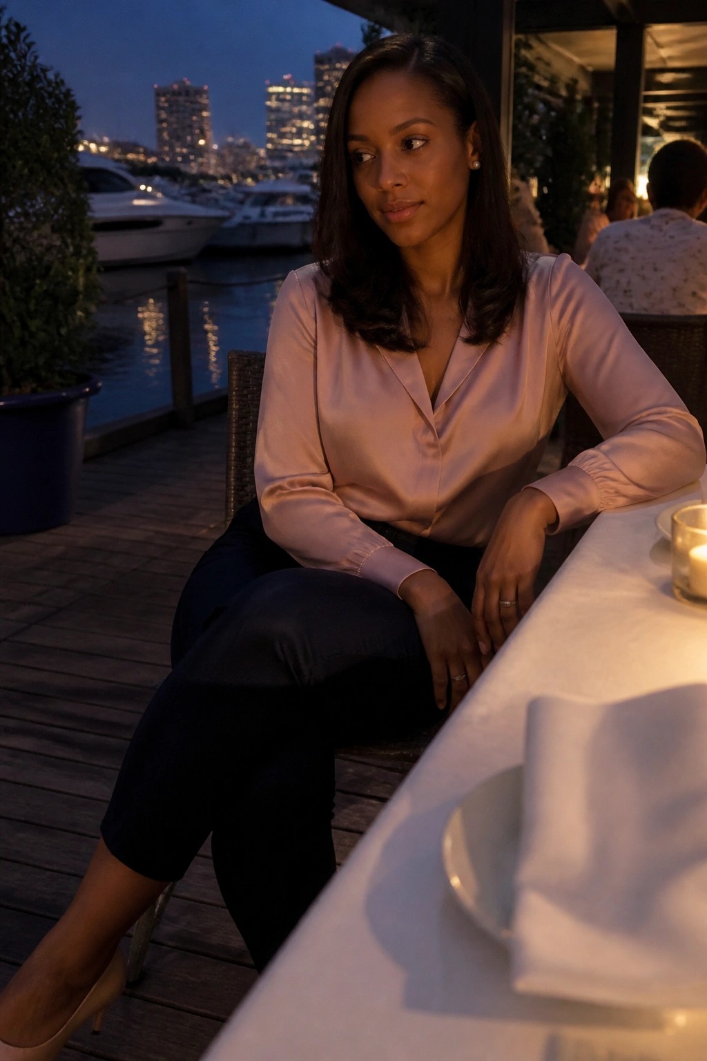 Woman in pink satin blouse at waterfront restaurant table at dusk.