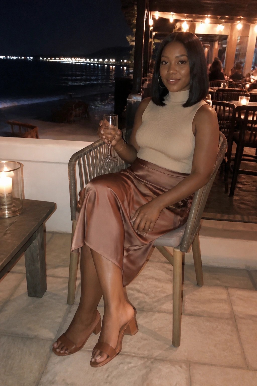 Woman in beige turtleneck and brown satin skirt holding wine glass at night oceanfront restaurant
