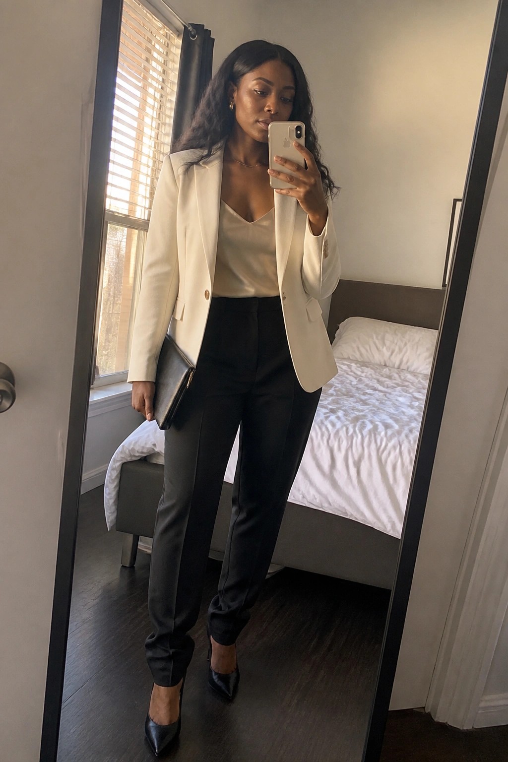 Woman in white blazer and black pants taking mirror selfie in bedroom.