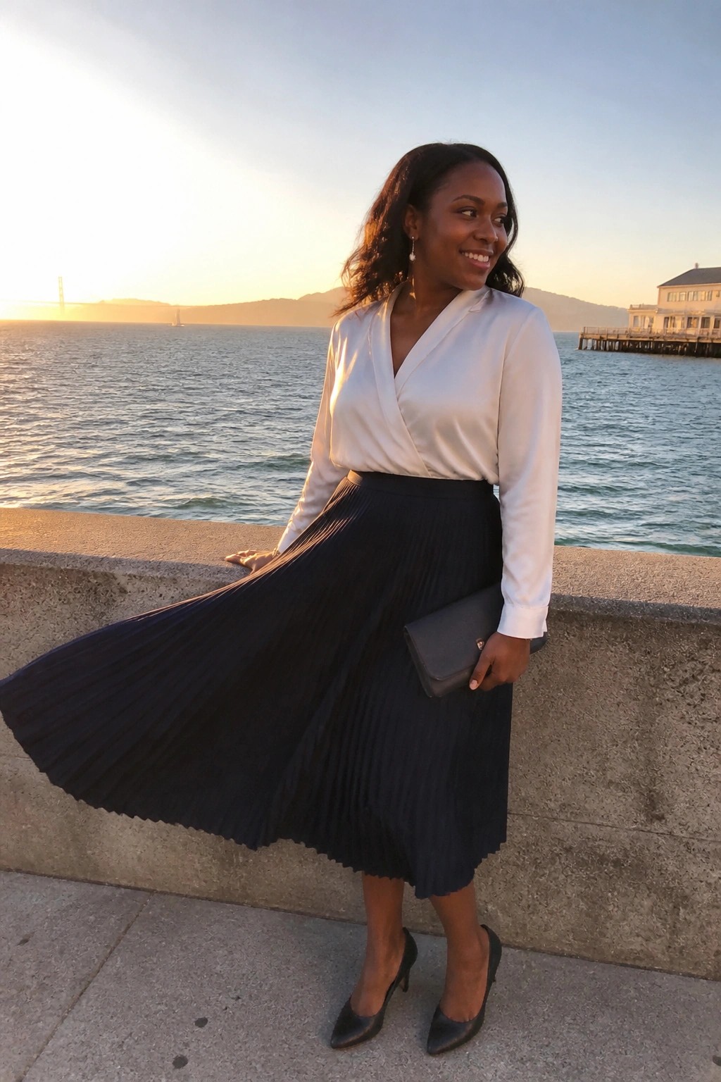 Woman in white blouse and flowing black pleated skirt by sunset waterfront