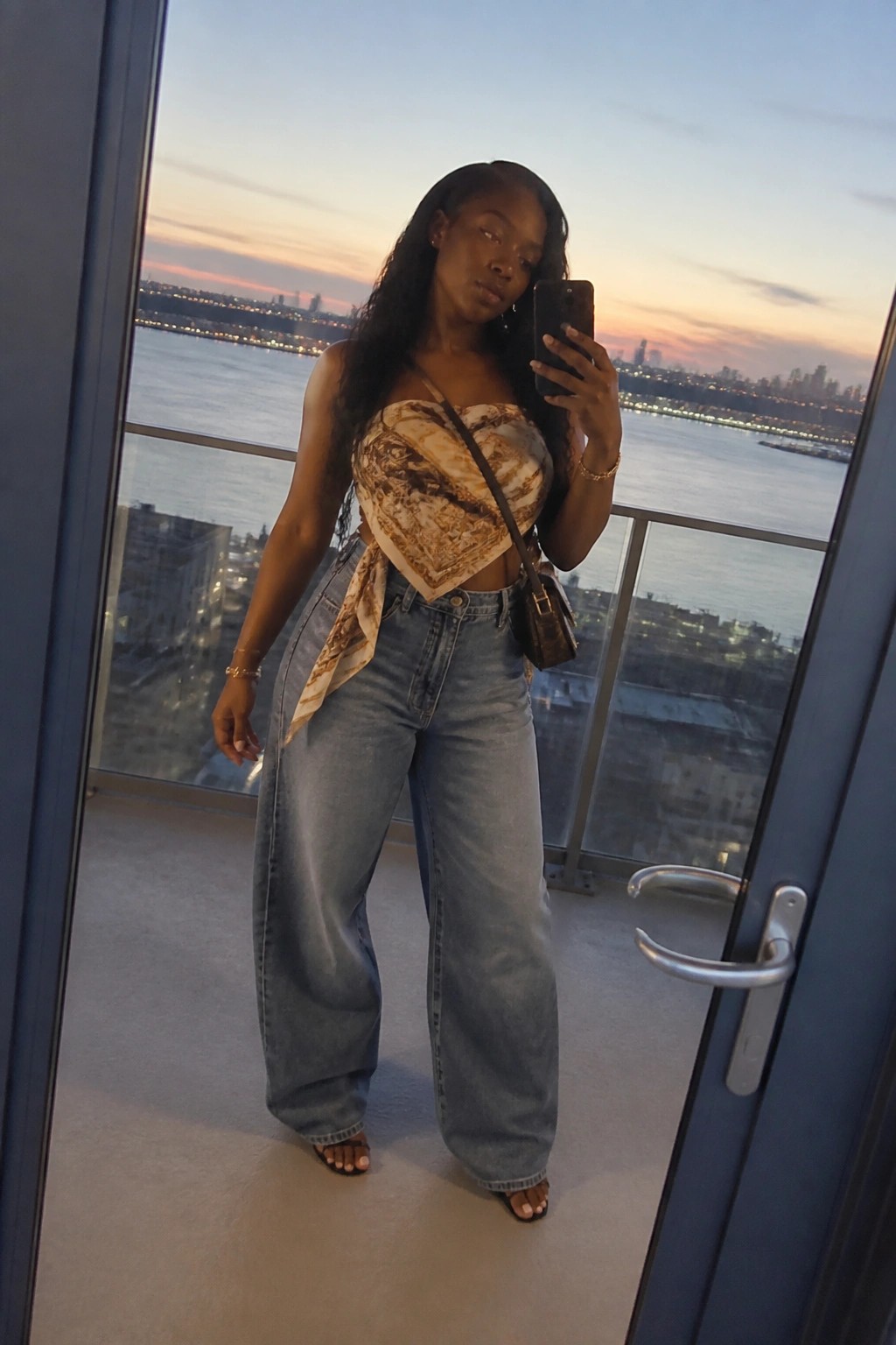 Woman in patterned crop top and wide-leg jeans taking selfie on sunset balcony