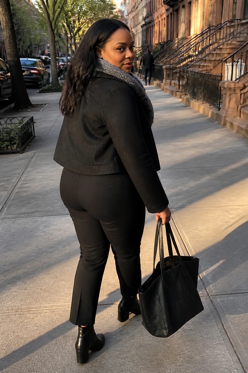 Woman in black jacket and pants with gray scarf holds tote on sidewalk