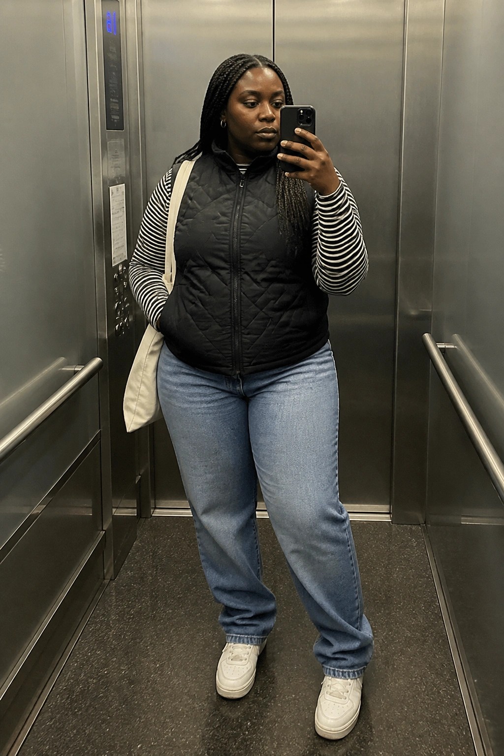 Woman taking selfie in elevator wearing black vest over striped shirt and jeans.